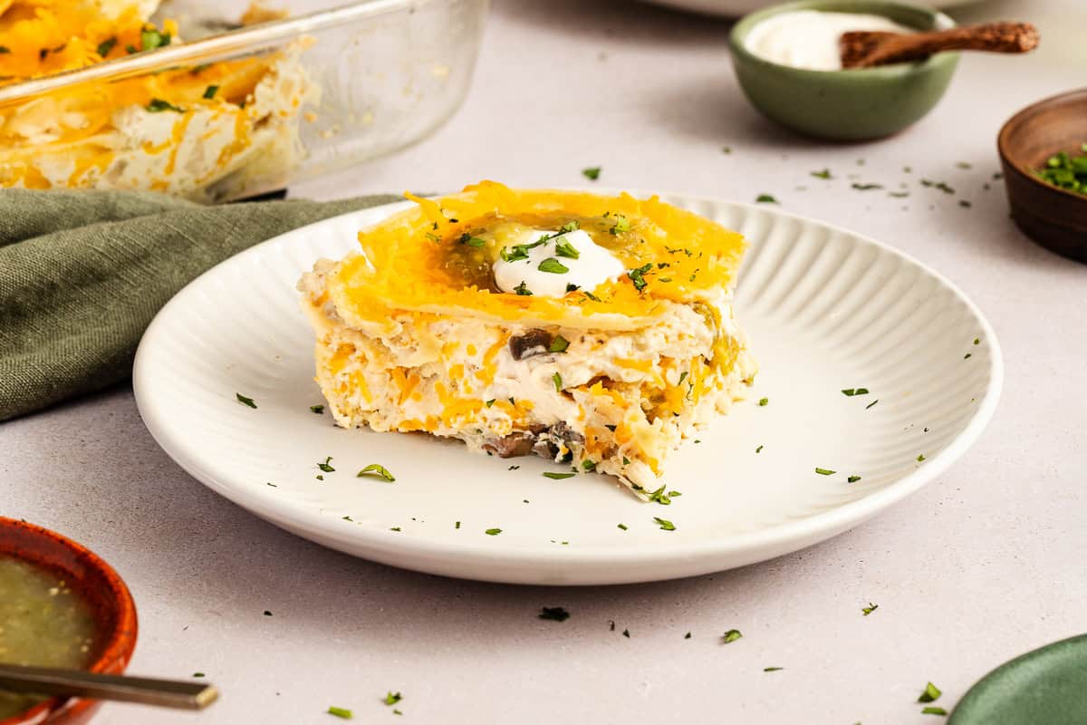 A serving of cheesy casserole with herbs and a dollop of sour cream on a white plate, surrounded by small bowls of condiments and a green napkin.
