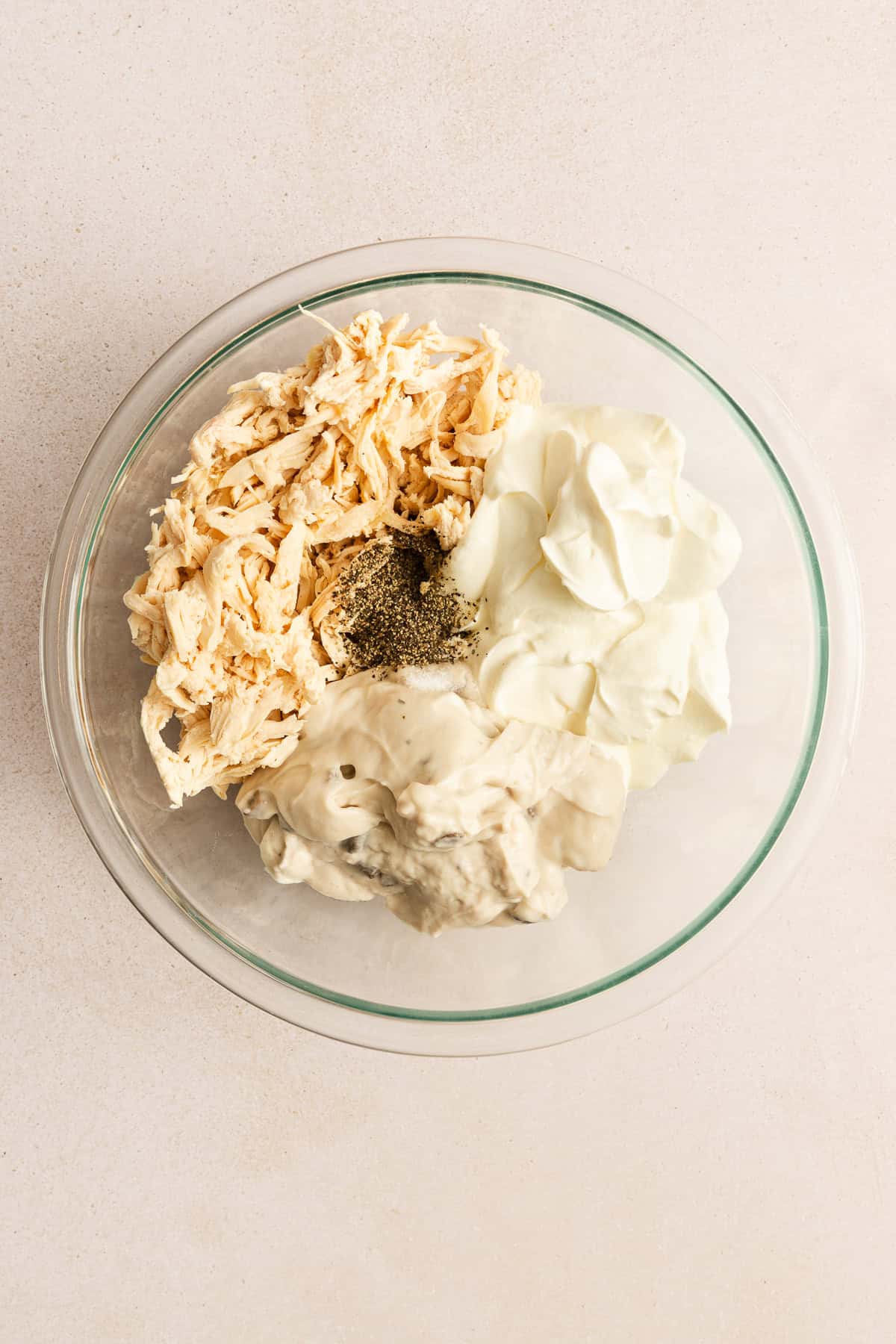 A glass bowl containing shredded chicken, sour cream, cream of mushroom soup, and black pepper on a light countertop.