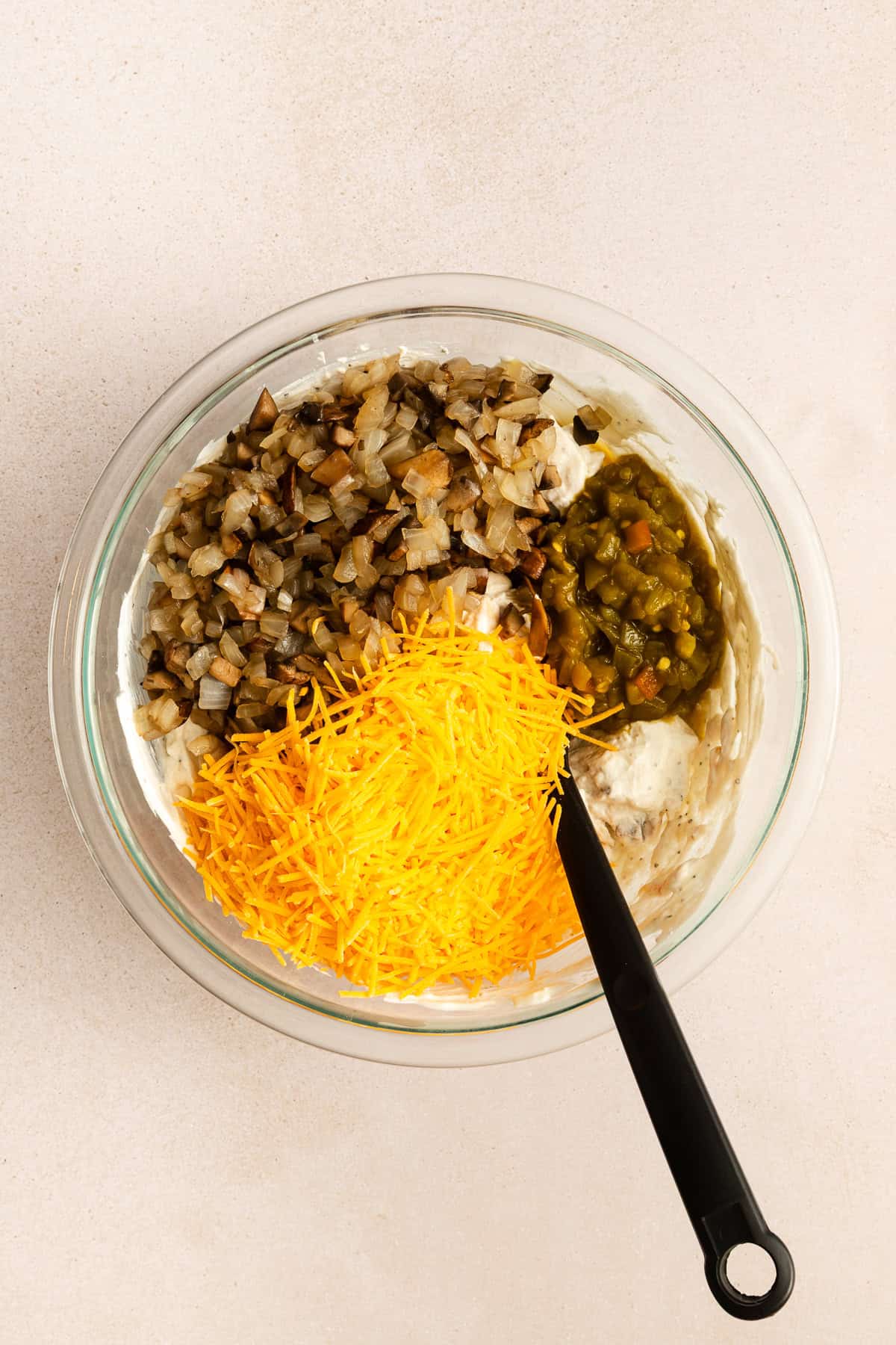 A glass bowl containing cream cheese, chopped saut&eacute;ed onions and mushrooms, chopped green chilies, and shredded cheddar cheese, with a black spoon inside.