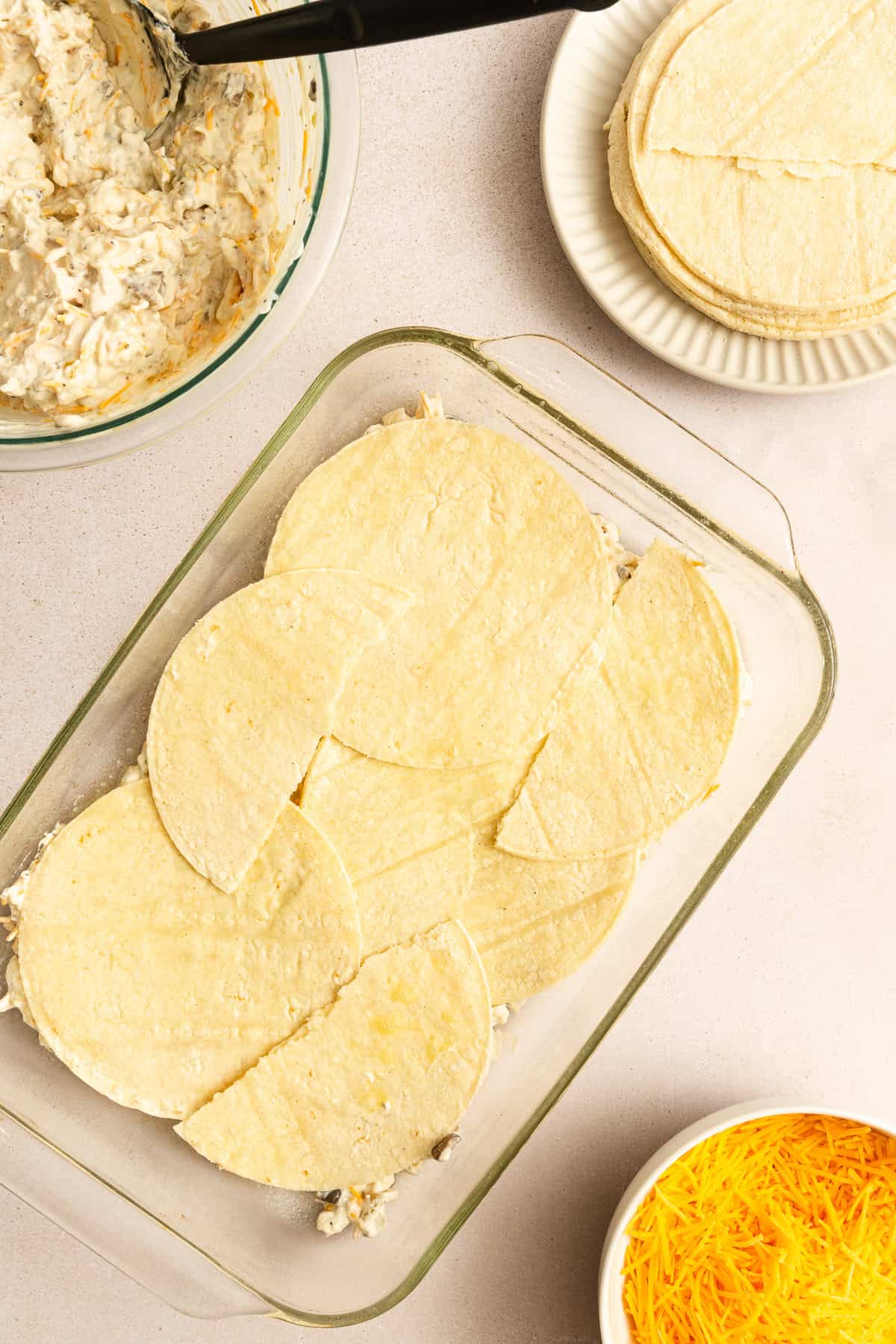 Glass baking dish with corn tortillas layered on top of a creamy mixture, surrounded by a bowl of shredded cheese, stacked tortillas, and a bowl of the same mixture with a spoon.
