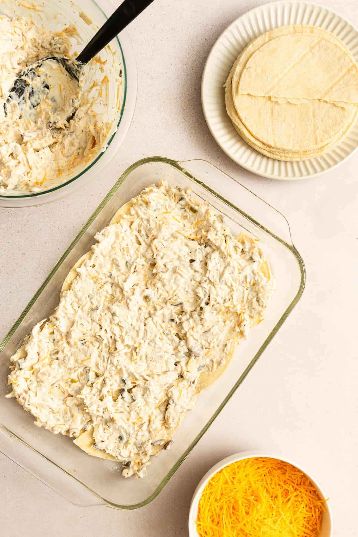 A glass baking dish with a creamy mixture spread over tortillas, next to a bowl of shredded cheese, a plate of tortillas, and a mixing bowl with more filling.