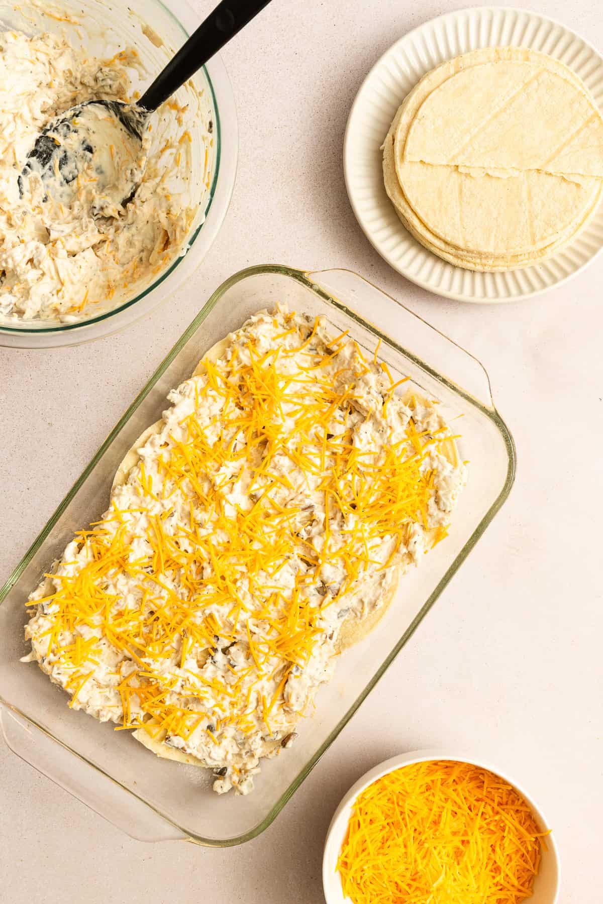 A glass baking dish with a layered casserole topped with shredded cheese, surrounded by a bowl of cheese, a plate of corn tortillas, and a bowl of creamy filling with a spatula.