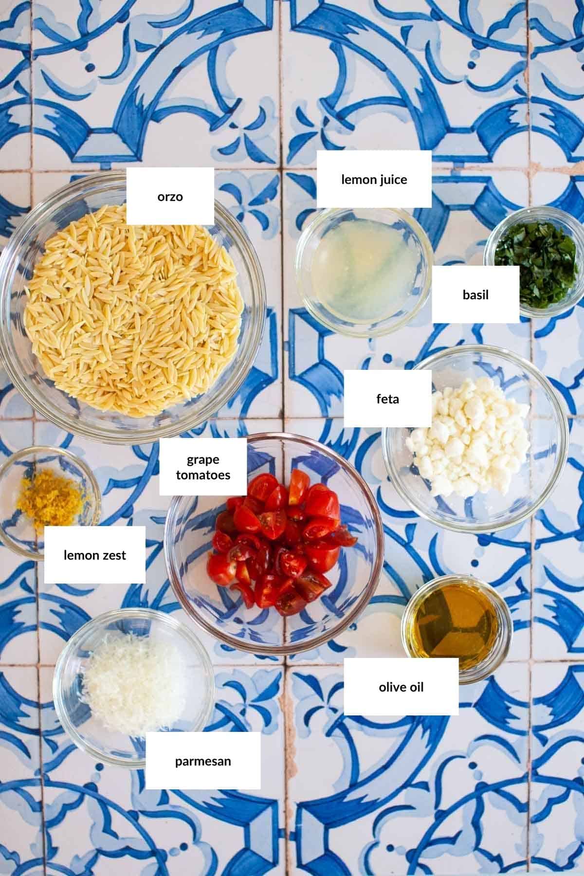 Top-down view of labeled ingredients for lemon orzo pasta salad in glass bowls on a blue and white tile surface: orzo, lemon juice, basil, feta, grape tomatoes, lemon zest, parmesan, and olive oil.