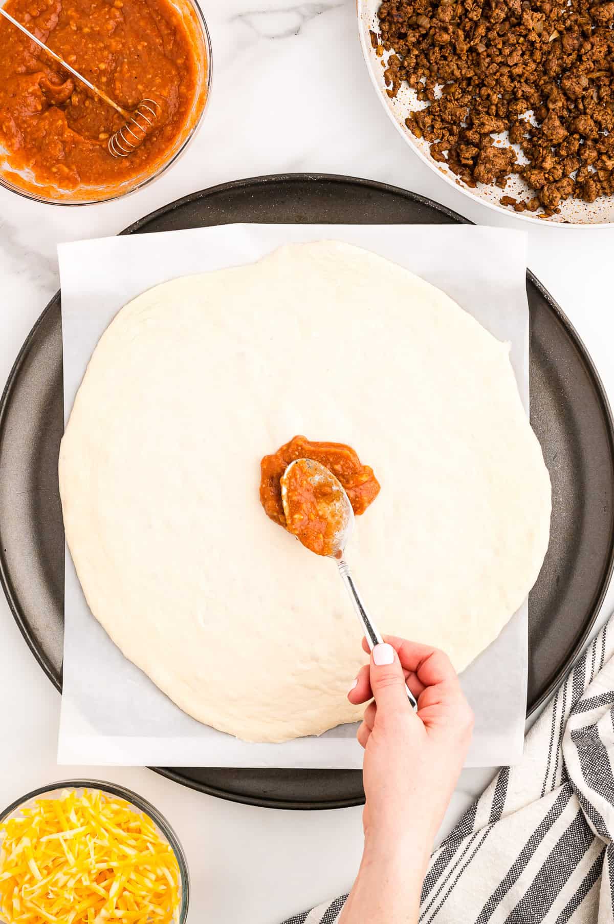 A hand spreading sauce onto a round sheet of pizza dough on a baking tray, with bowls of shredded cheese, ground meat, and extra sauce nearby.