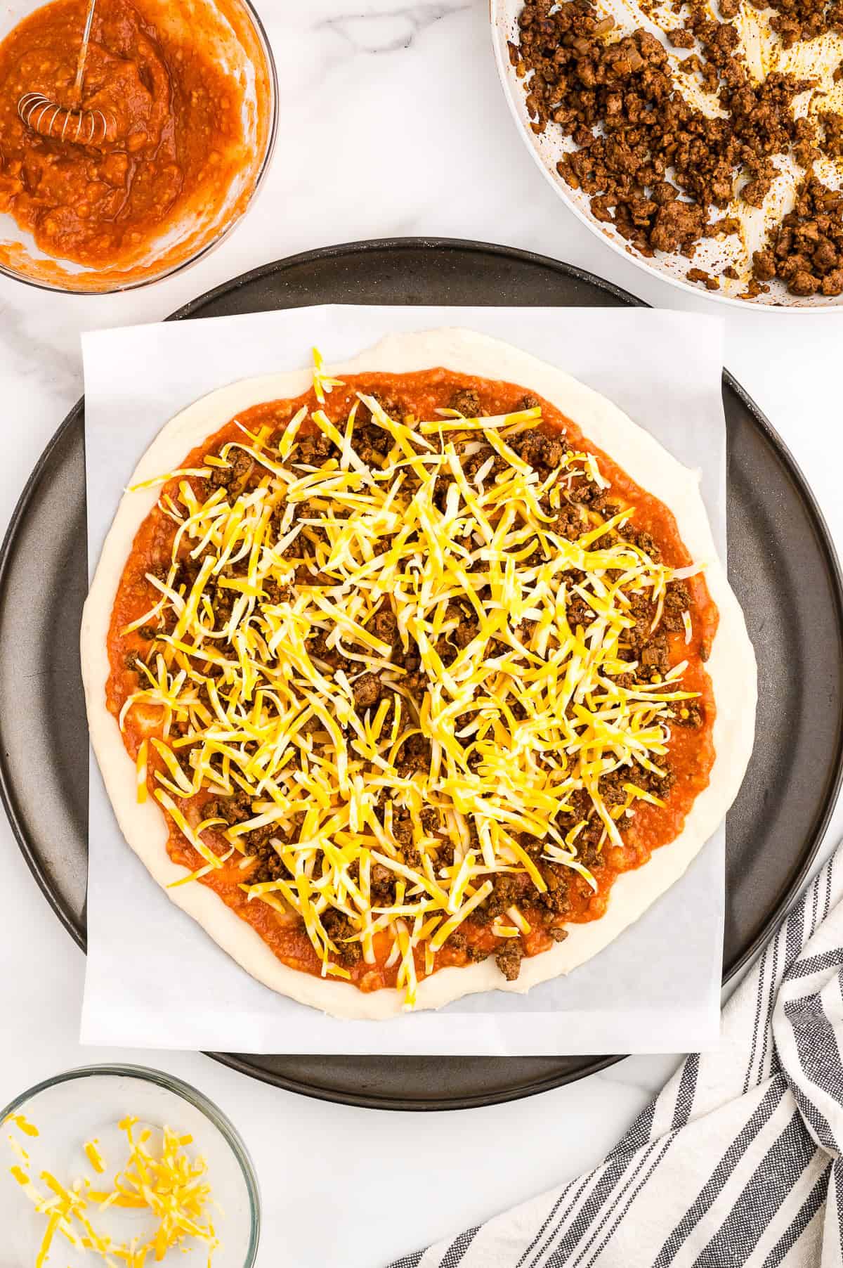 Unbaked pizza on parchment paper topped with tomato sauce, ground meat, and shredded cheese, with extra sauce, cheese, and meat on the side.