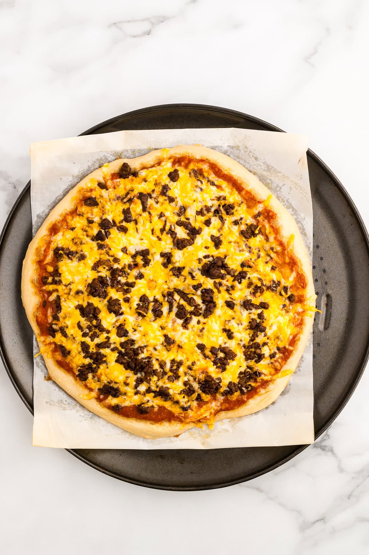 A baked pizza topped with ground beef and melted cheddar cheese sits on parchment paper atop a round, dark baking tray on a marble surface.