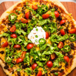 A taco-inspired pizza topped with ground beef, cheddar cheese, lettuce, cherry tomatoes, jalapeño slices, and a dollop of sour cream on a wooden board.