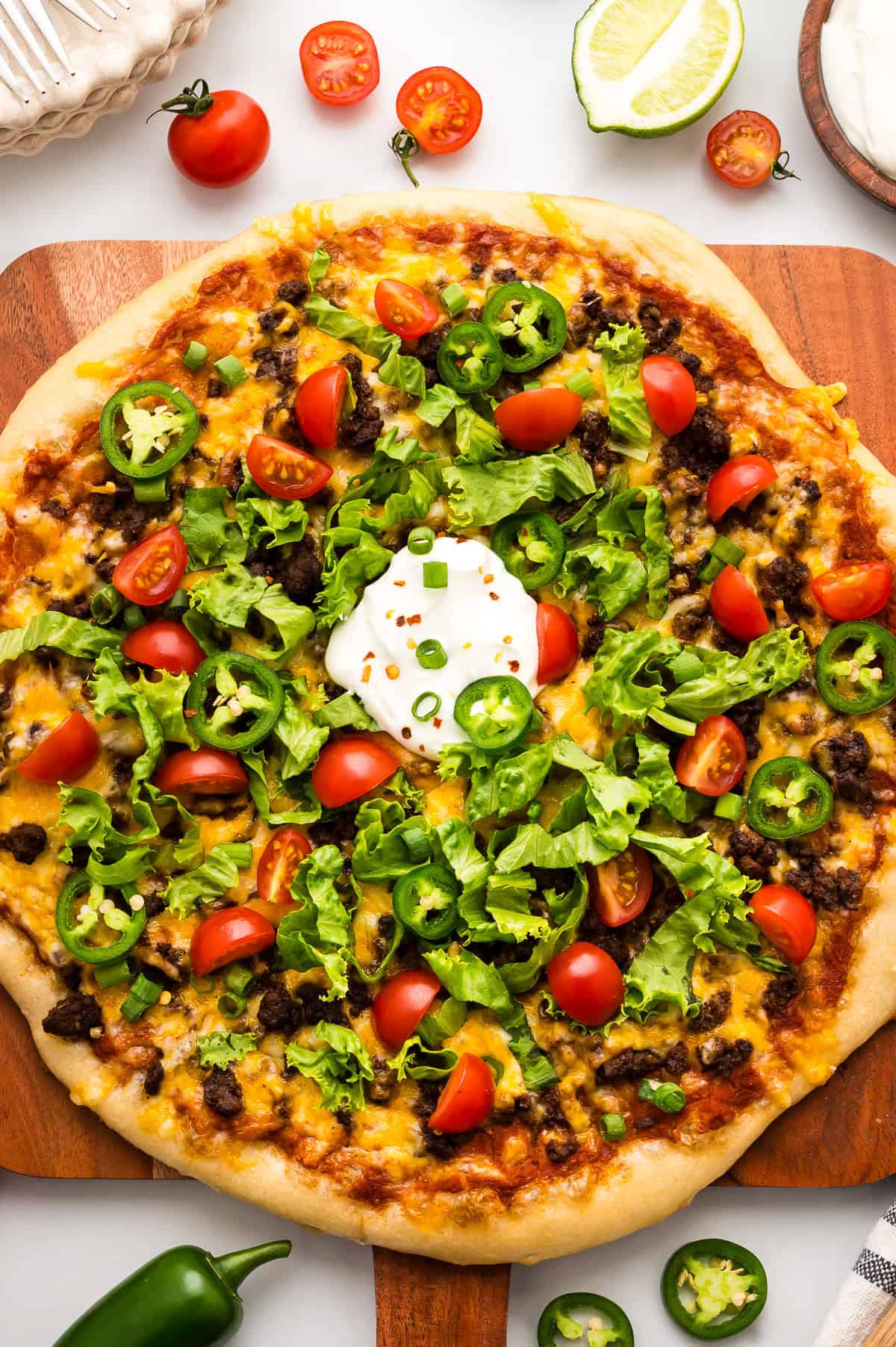 A taco-inspired pizza topped with ground beef, cheddar cheese, lettuce, cherry tomatoes, jalape&ntilde;o slices, and a dollop of sour cream on a wooden board.