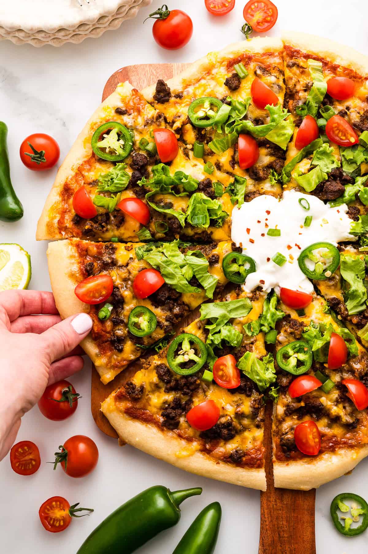 A hand takes a slice of taco pizza topped with ground beef, lettuce, cherry tomatoes, jalape&ntilde;os, cheese, and a dollop of sour cream, surrounded by fresh vegetables.