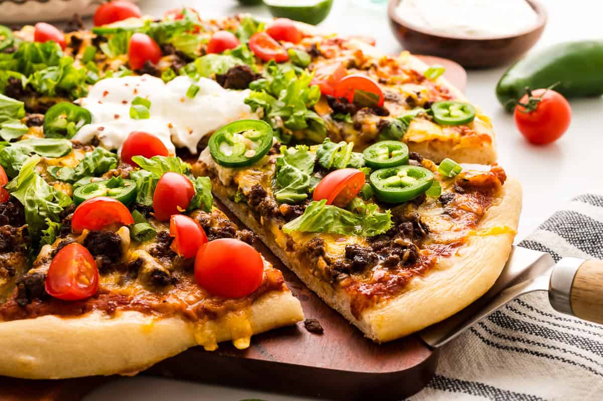 A taco pizza topped with ground beef, cheese, lettuce, cherry tomatoes, jalape&ntilde;o slices, and a dollop of sour cream, with one slice being served.