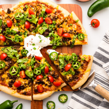 A taco-inspired pizza topped with ground beef, cheese, lettuce, cherry tomatoes, jalapeños, and sour cream, surrounded by fresh ingredients on a white surface.
