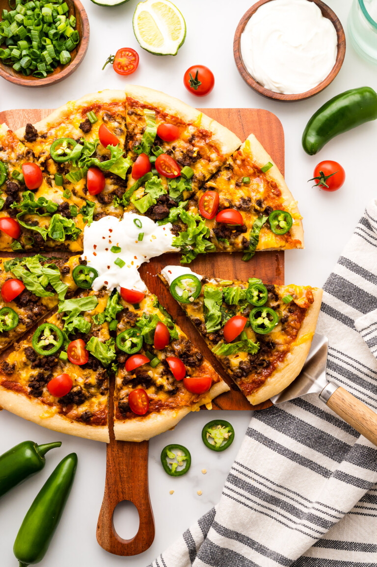 A taco-inspired pizza topped with ground beef, cheese, lettuce, cherry tomatoes, jalapeños, and sour cream, surrounded by fresh ingredients on a white surface.