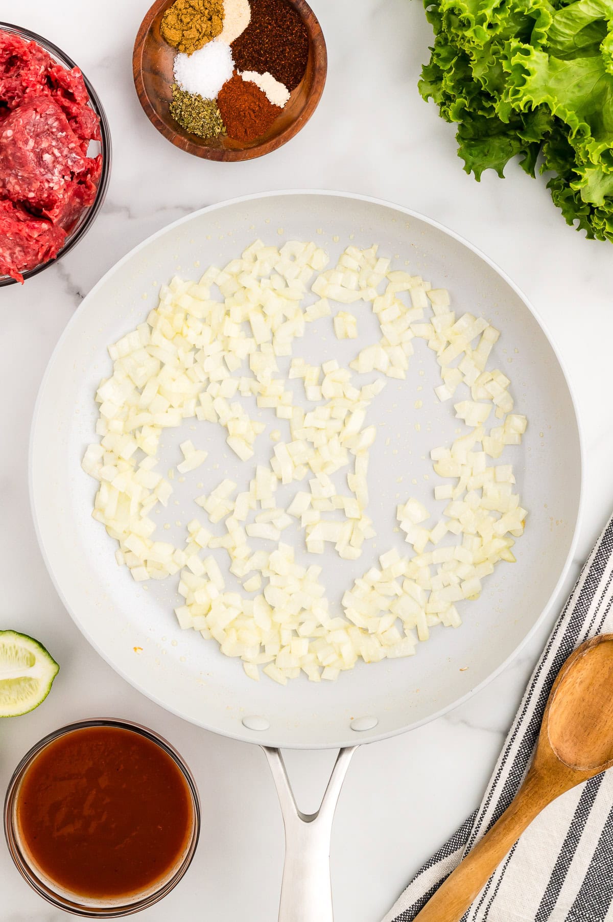 Diced onions saut&eacute;ing in a white skillet, surrounded by raw ground beef, spices, green lettuce, sauce, a lime wedge, and a wooden spoon.