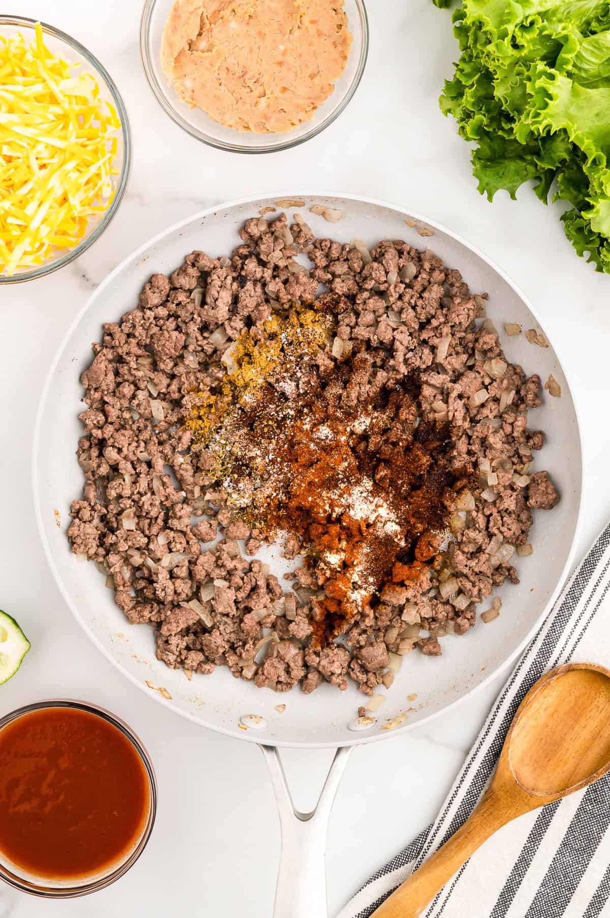 Ground beef and diced onions cooking in a skillet with various seasonings on top, surrounded by shredded cheese, refried beans, lettuce, enchilada sauce, and a wooden spoon.