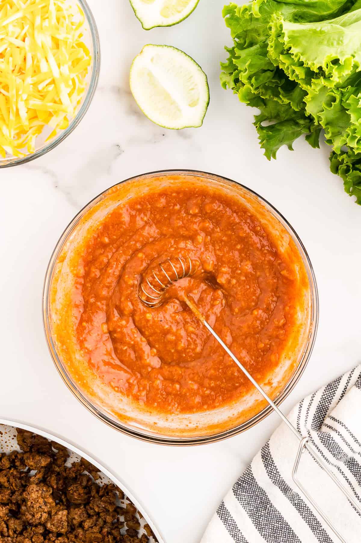 A bowl of red sauce with a whisk, surrounded by shredded cheese, lettuce, lime halves, cooked ground meat, and a striped towel on a white surface.