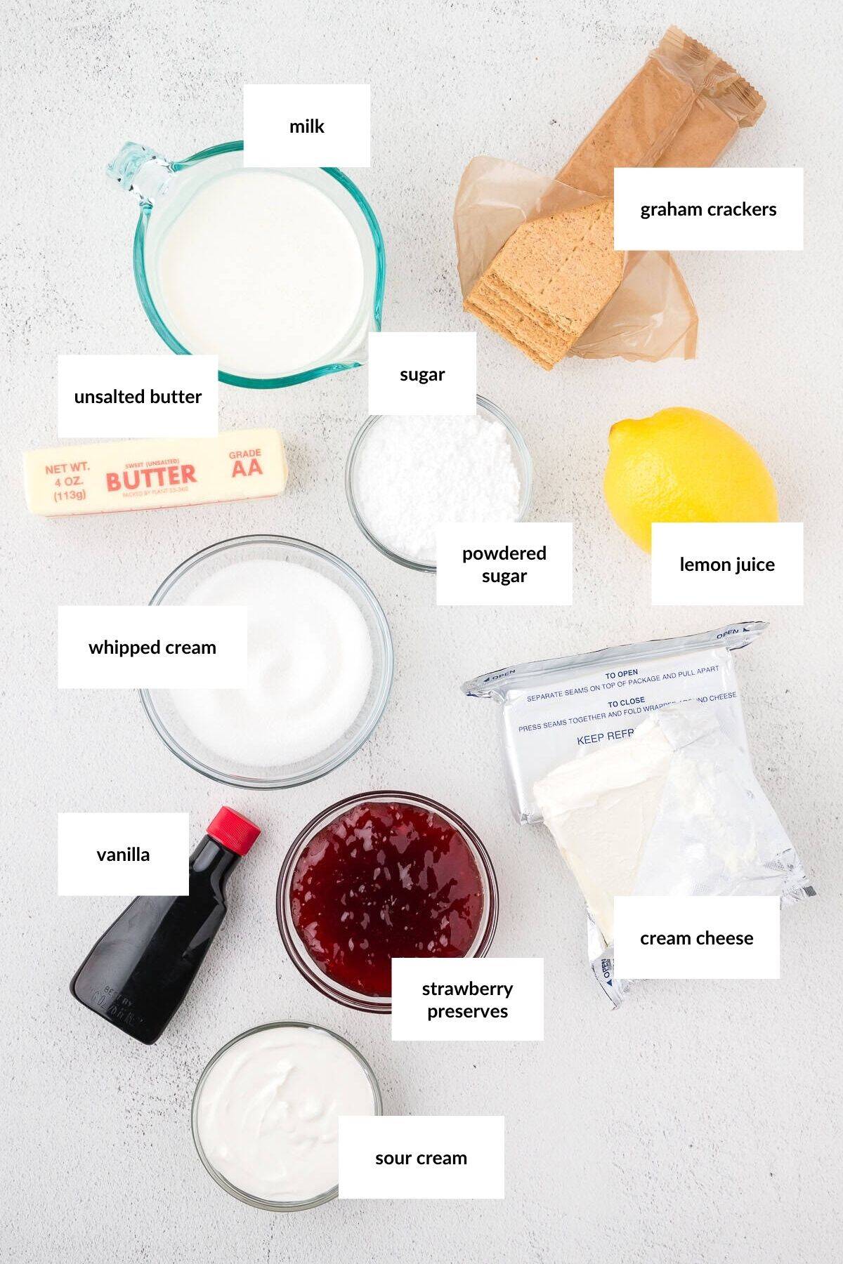 Ingredients for a dessert recipe arranged on a white surface, each labeled: milk, graham crackers, unsalted butter, sugar, powdered sugar, lemon juice, whipped cream, vanilla, strawberry preserves, cream cheese, sour cream.