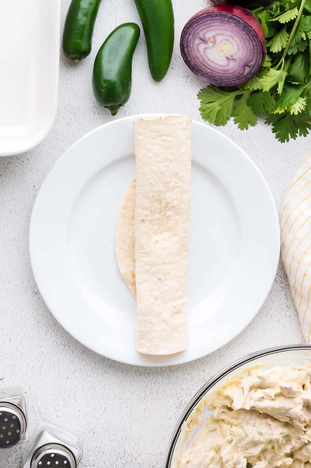 A rolled tortilla is placed on a white plate, surrounded by jalape&ntilde;os, sliced red onion, cilantro, and a bowl of creamy filling.