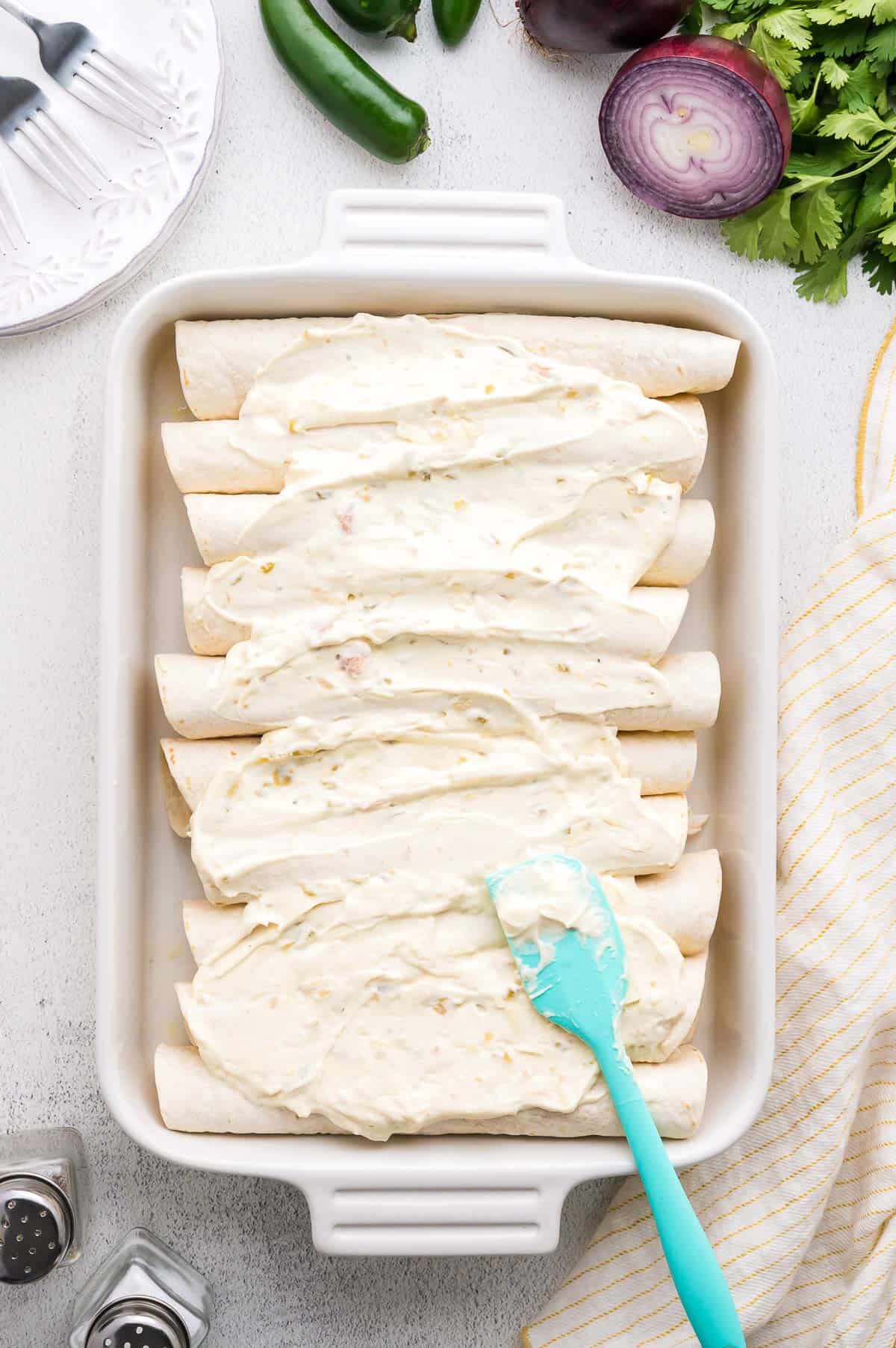 A baking dish filled with rolled tortillas covered in a creamy white sauce, with a blue spatula spreading the sauce; sliced vegetables and seasonings are nearby.