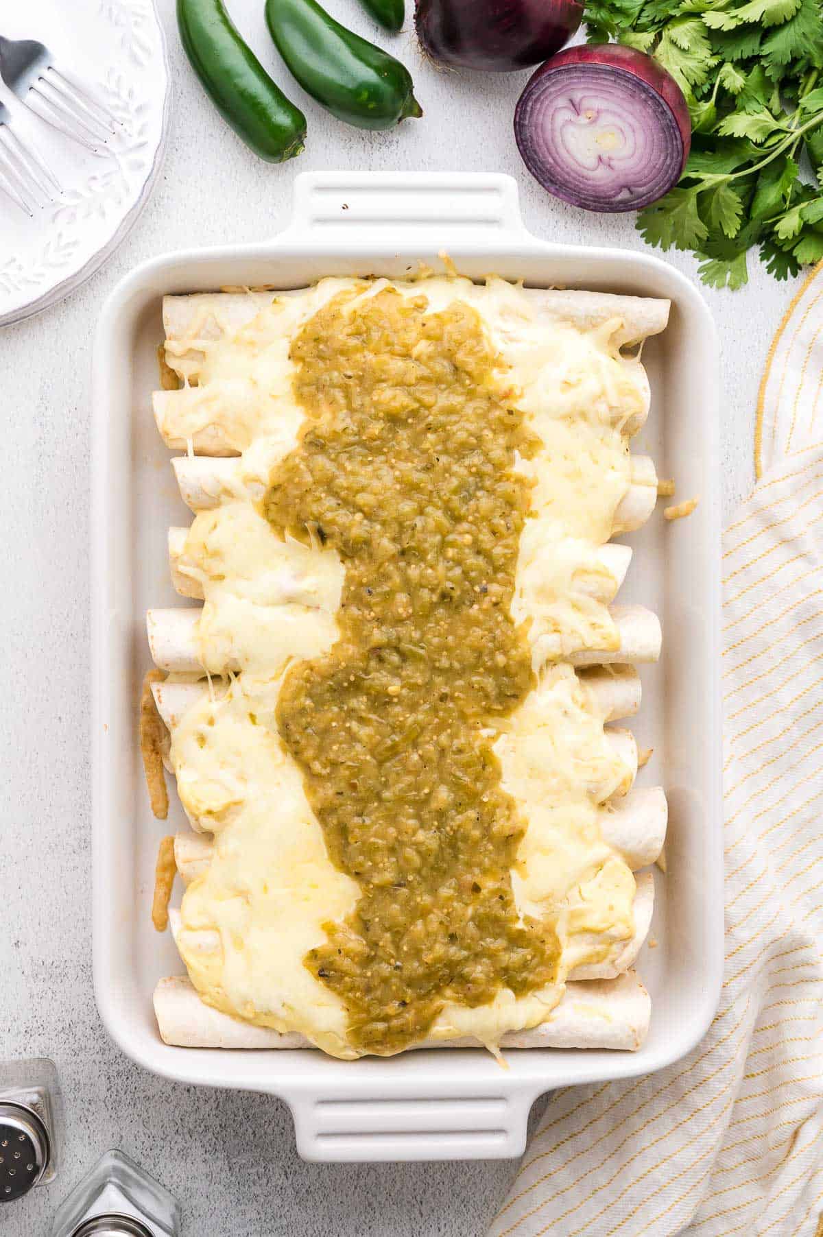 A white baking dish with six cheese-covered enchiladas topped with green salsa. Fresh cilantro, jalape&ntilde;os, and a red onion are arranged nearby.