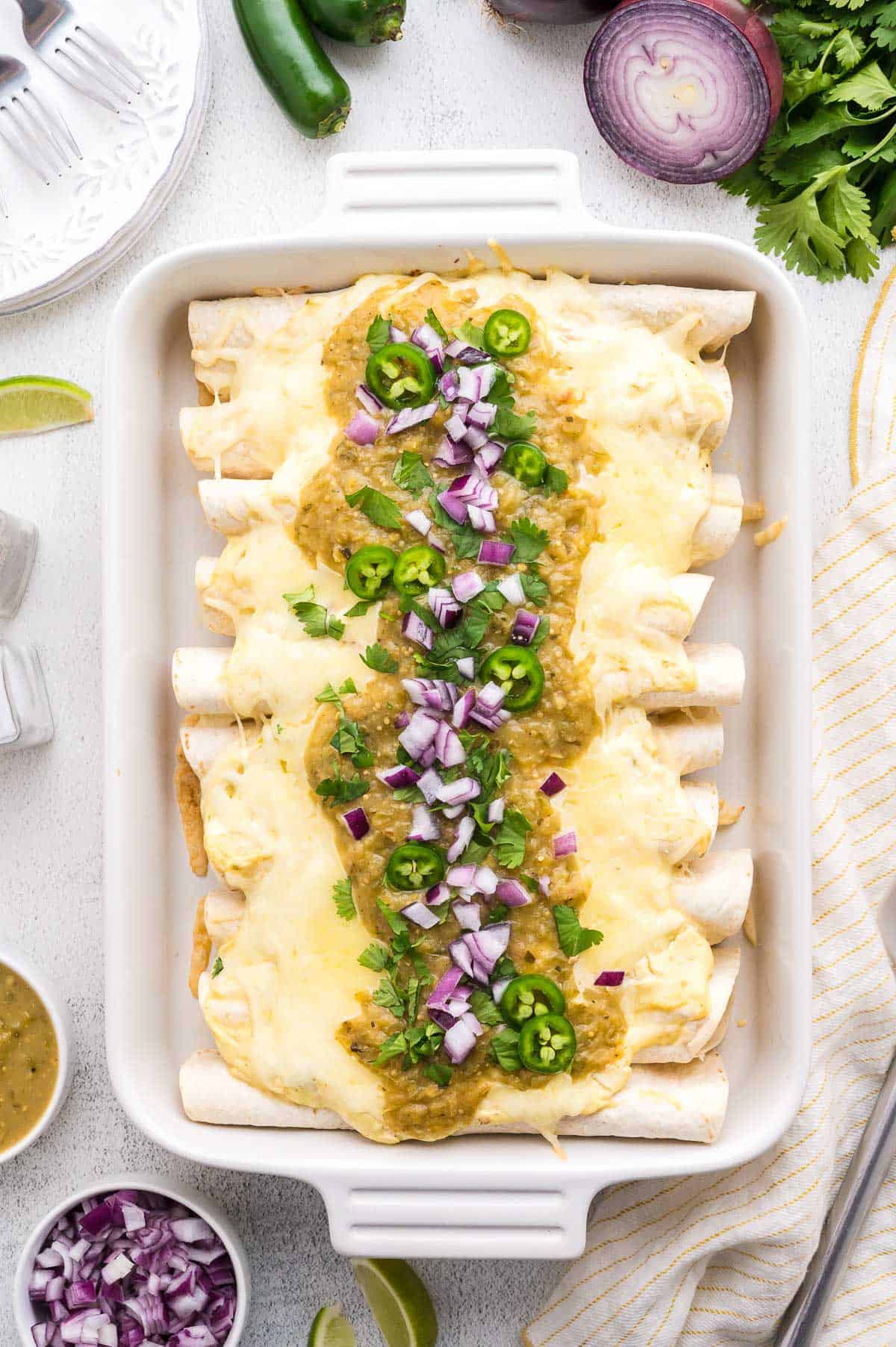 A baking dish filled with cheese-covered sour cream enchiladas, topped with green salsa, sliced jalape&ntilde;os, and chopped red onions.