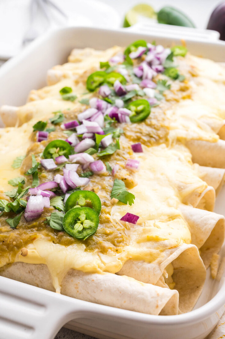 A baking dish filled with enchiladas topped with melted cheese, green sauce, sliced jalape&ntilde;os, chopped red onion, and cilantro.