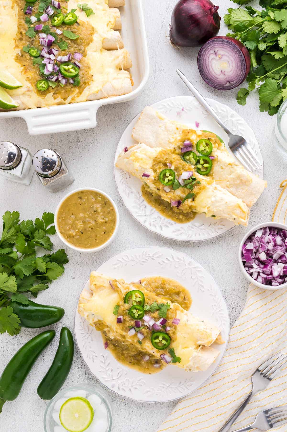 Two plates of enchiladas topped with green sauce, sliced jalape&ntilde;os, and chopped red onions. Additional garnishes and ingredients are arranged around the dishes.