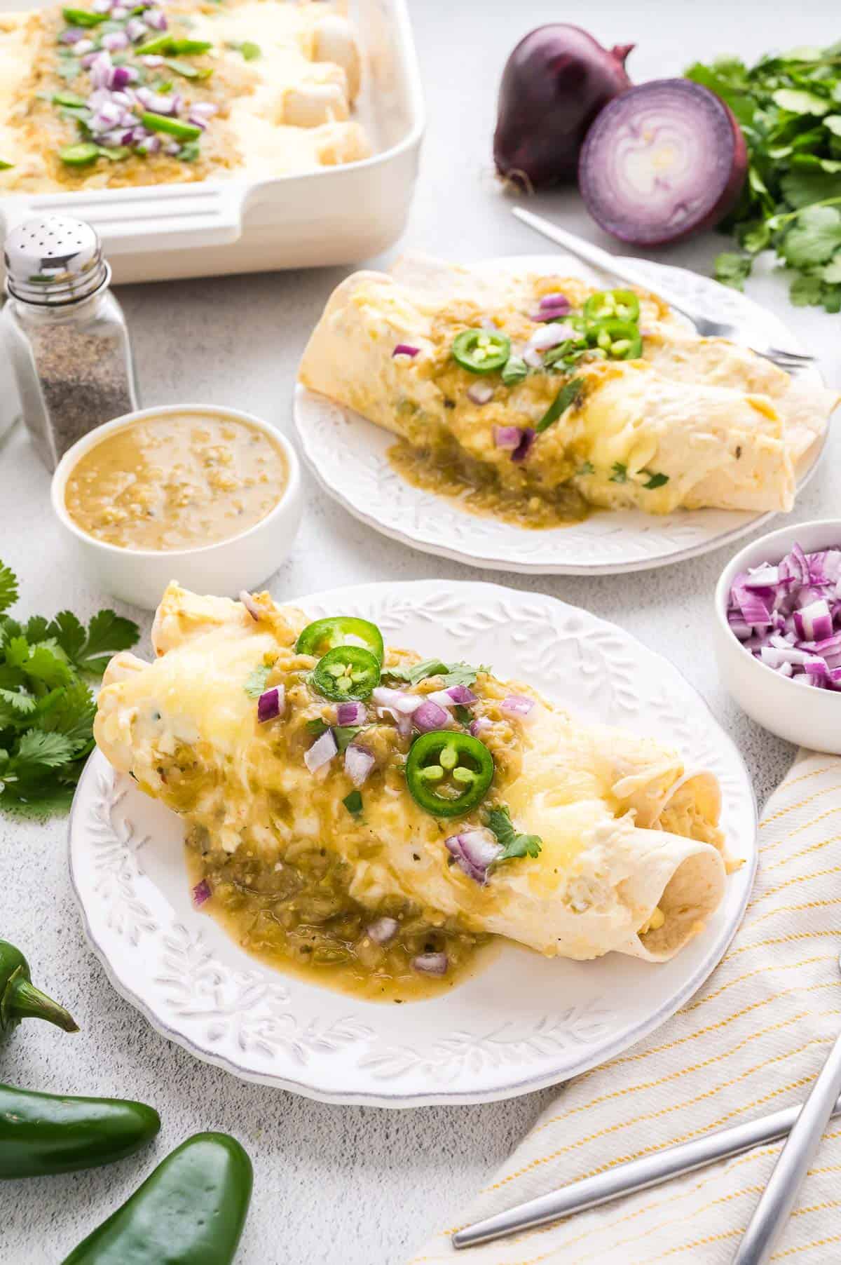 Two plates of enchiladas topped with green salsa, sliced jalape&ntilde;os, and chopped red onion, surrounded by ingredients like cilantro, jalape&ntilde;os, and a bowl of salsa.
