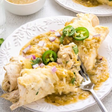 A plate of chicken enchiladas topped with melted cheese, green salsa, sliced jalapeños, and diced red onions, with a fork and a bowl of salsa on the side.