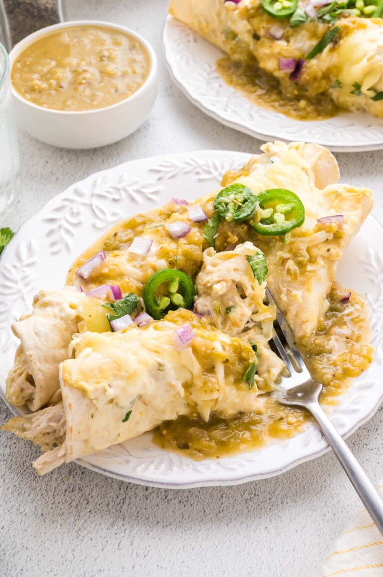 A plate of chicken enchiladas topped with melted cheese, green salsa, sliced jalapeños, and diced red onions, with a fork and a bowl of salsa on the side.