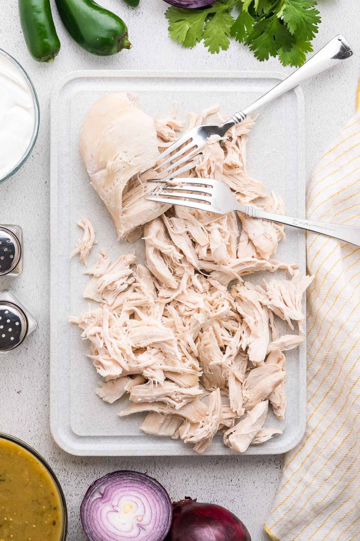 Two forks shred cooked chicken breast on a white cutting board, surrounded by ingredients like jalape&ntilde;os, cilantro, onions, sour cream, and sauce bowls.