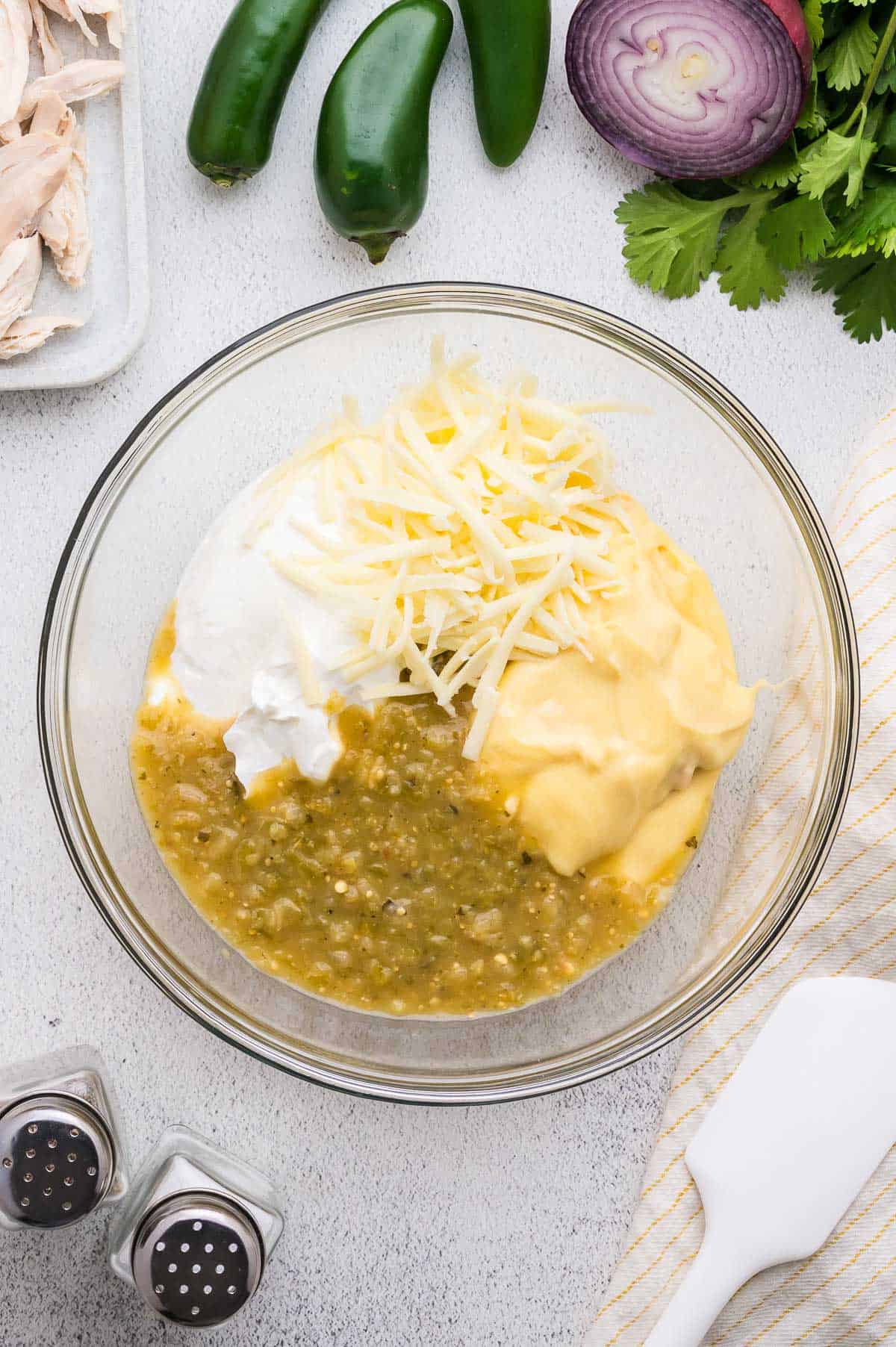 A glass bowl contains salsa verde, shredded cheese, sour cream, and creamy soup mix. Sliced red onion, jalape&ntilde;os, cilantro, and shredded chicken are nearby on the countertop.
