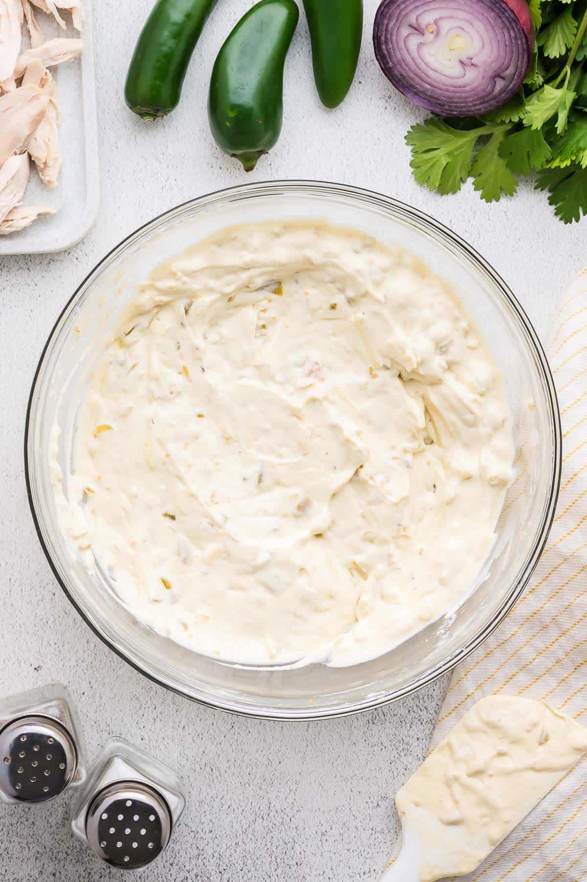 A glass bowl filled with creamy dip mixture surrounded by jalape&ntilde;os, red onion, cilantro, shredded chicken, and a striped towel.