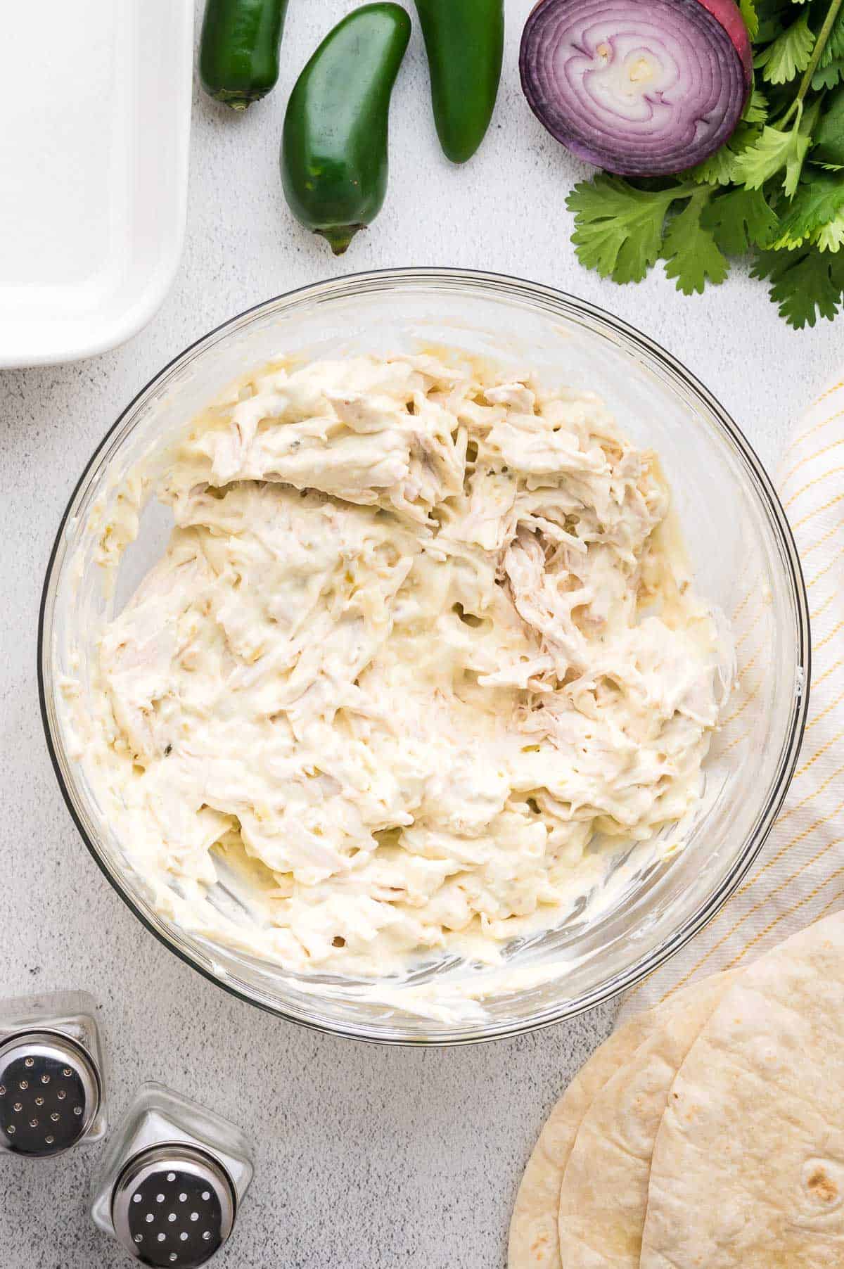 A glass bowl filled with shredded chicken mixed with a creamy white sauce, surrounded by tortillas, jalape&ntilde;os, red onion, cilantro, and salt and pepper shakers on a countertop.