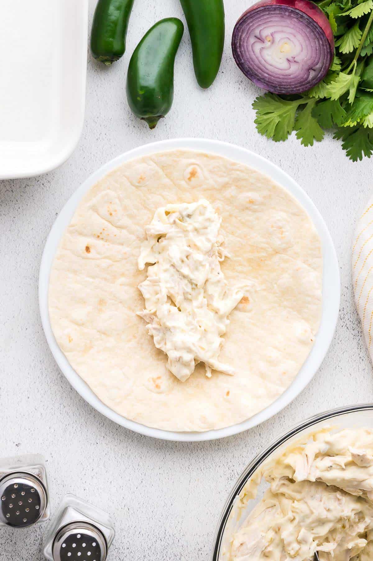A flour tortilla topped with a creamy chicken mixture sits on a white plate, surrounded by jalape&ntilde;os, red onion, cilantro, and salt and pepper shakers.