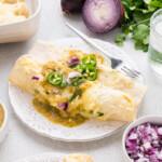 A plate of enchiladas topped with green salsa, sliced jalapeños, and chopped red onion, next to a glass of water with lime and bowls of salsa and diced onions.