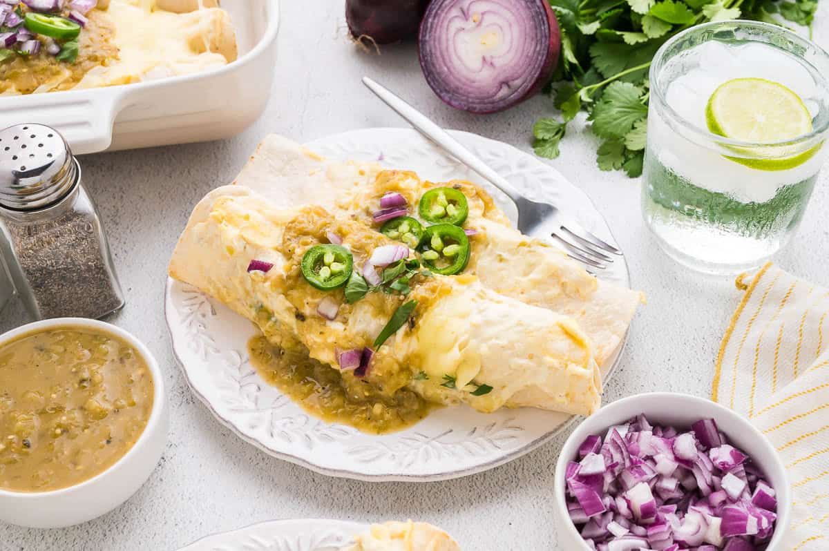 A plate of enchiladas topped with green salsa, sliced jalape&ntilde;os, and chopped red onion, next to a glass of water with lime and bowls of salsa and diced onions.