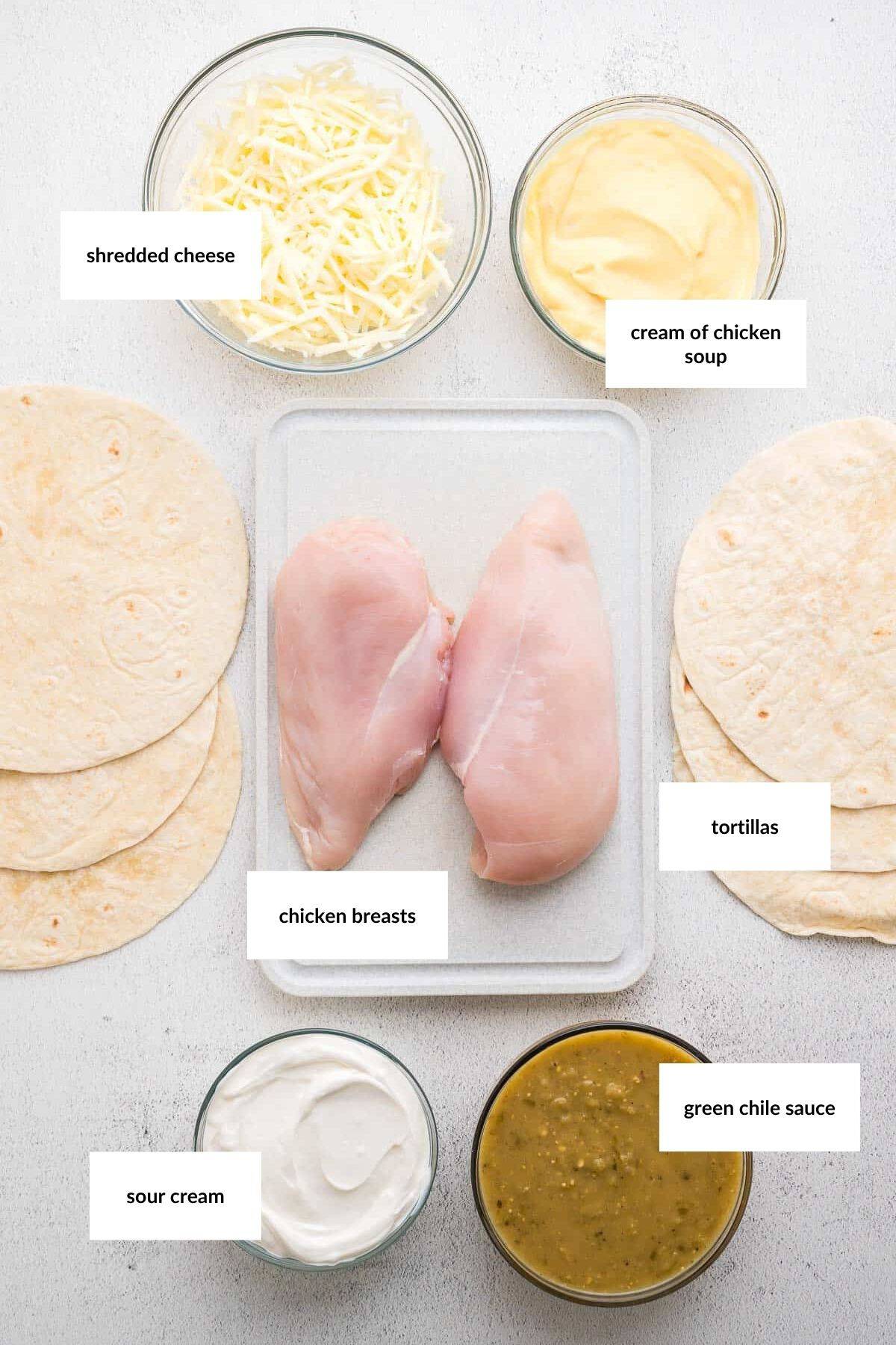 Ingredients for a chicken dish displayed: raw chicken breasts, shredded cheese, cream of chicken soup, tortillas, sour cream, and green chile sauce arranged on a white surface.