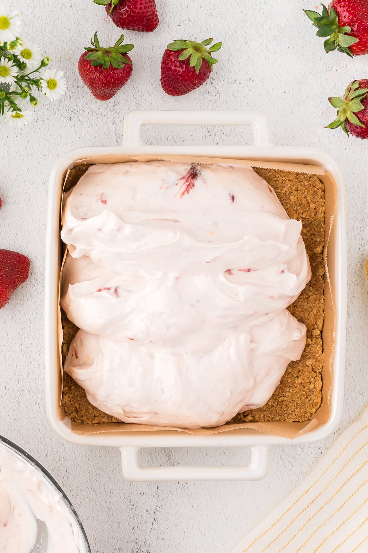 A square baking dish with a graham cracker crust topped with a layer of strawberry cream mixture, surrounded by fresh strawberries.