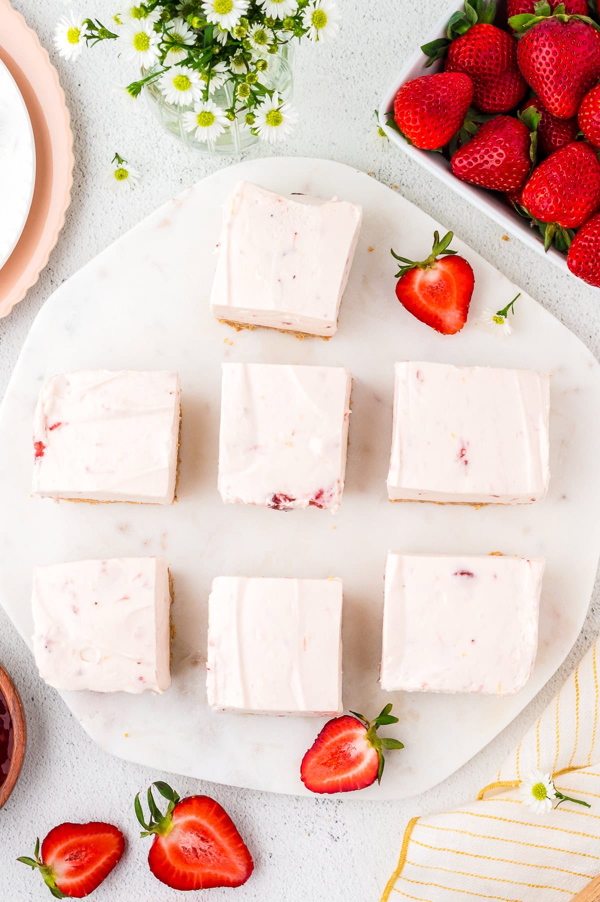 Six strawberry cheesecake bars on a round marble board, surrounded by fresh strawberries, a small bouquet of flowers, and a striped napkin.