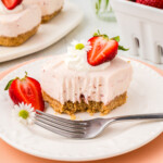 A slice of strawberry cheesecake bar with a bite taken out, topped with whipped cream, a halved strawberry, and a small flower on a white plate with a fork. Other bars and strawberries in the background.
