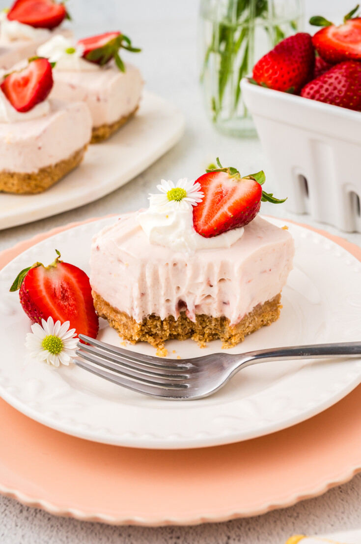 A slice of strawberry cheesecake bar with a bite taken out, topped with whipped cream, a halved strawberry, and a small flower on a white plate with a fork. Other bars and strawberries in the background.
