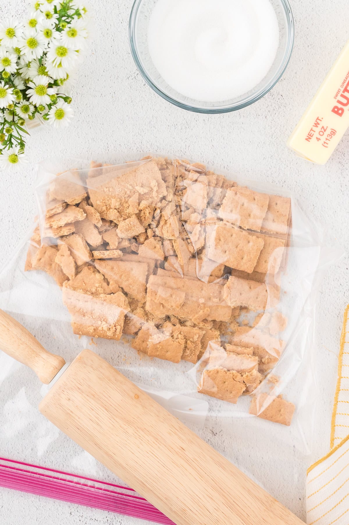 A rolling pin, a plastic bag with crushed graham crackers, a stick of butter, a bowl of sugar, flowers, and a striped towel on a light surface.