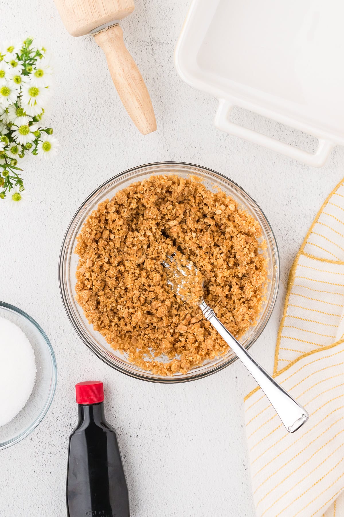 A glass bowl of crumbly graham cracker mixture with a fork, surrounded by a rolling pin, baking dish, striped towel, sugar, vanilla extract, and flowers on a white surface.