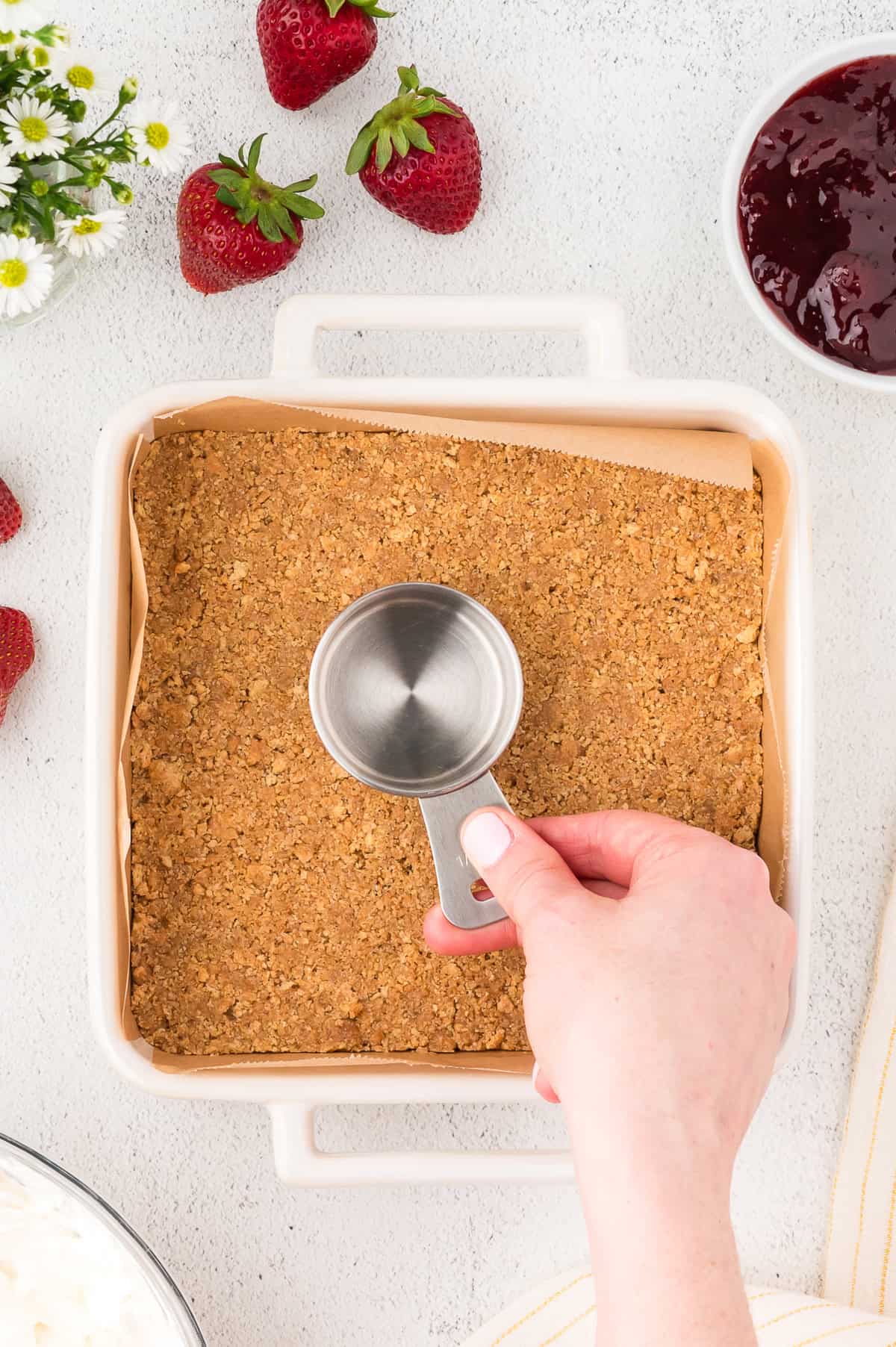 A hand presses a crumb crust into a square baking dish with a metal measuring cup. Strawberries, jam, and flowers are arranged around the dish on a white surface.