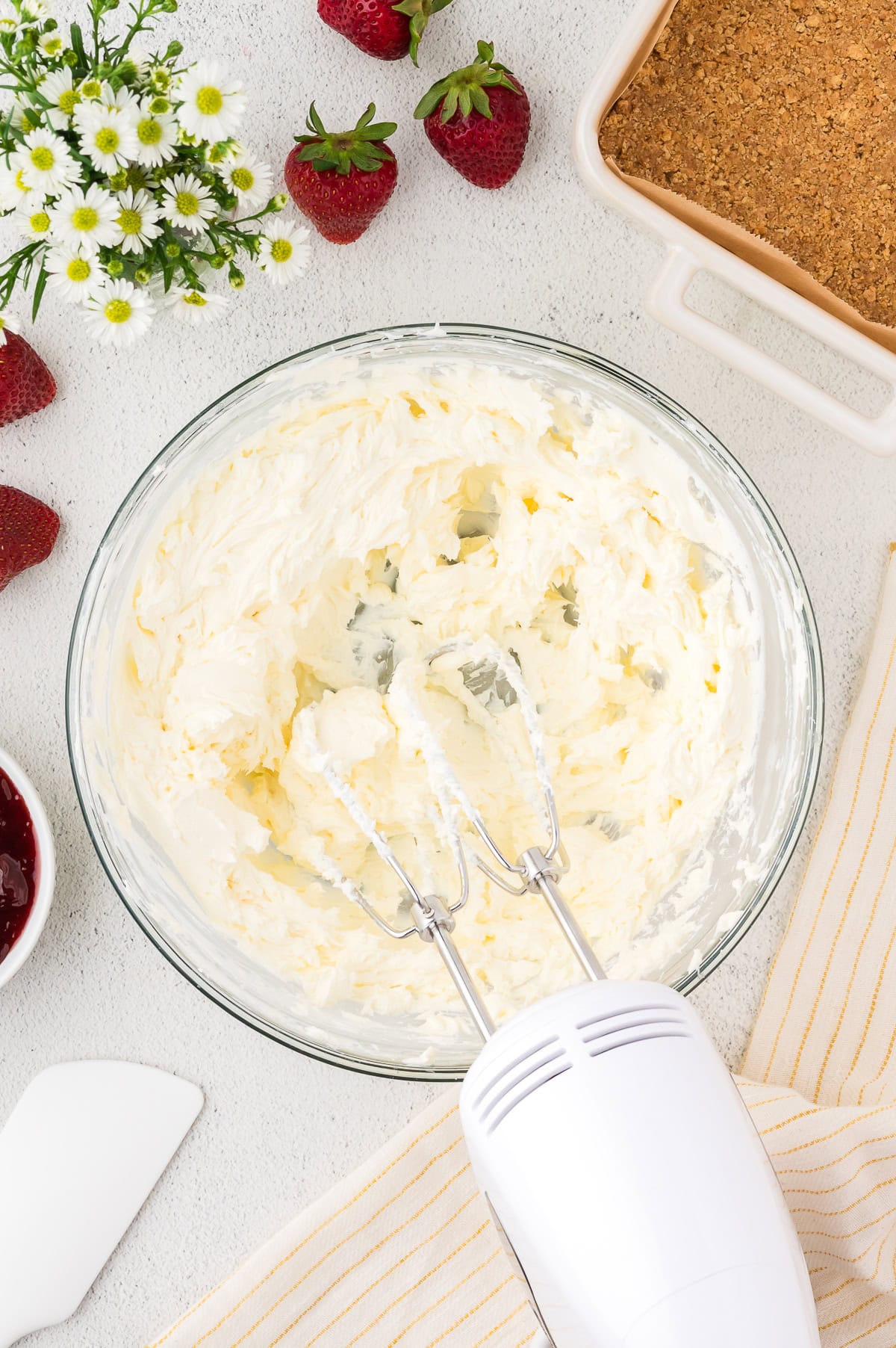 A bowl of whipped cream cheese mixture with an electric hand mixer, surrounded by strawberries, flowers, a baking dish with crust, and kitchen utensils on a countertop.