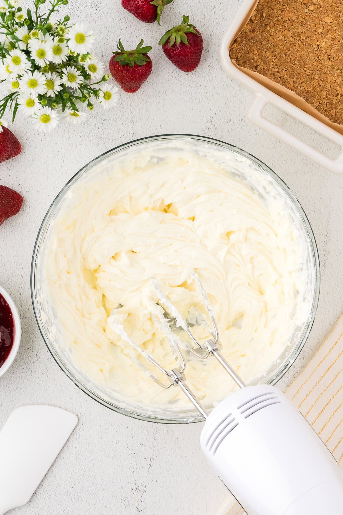 A hand mixer with beaters rests in a glass bowl of whipped cream cheese mixture, surrounded by strawberries, flowers, and a baking dish with a crumb crust.