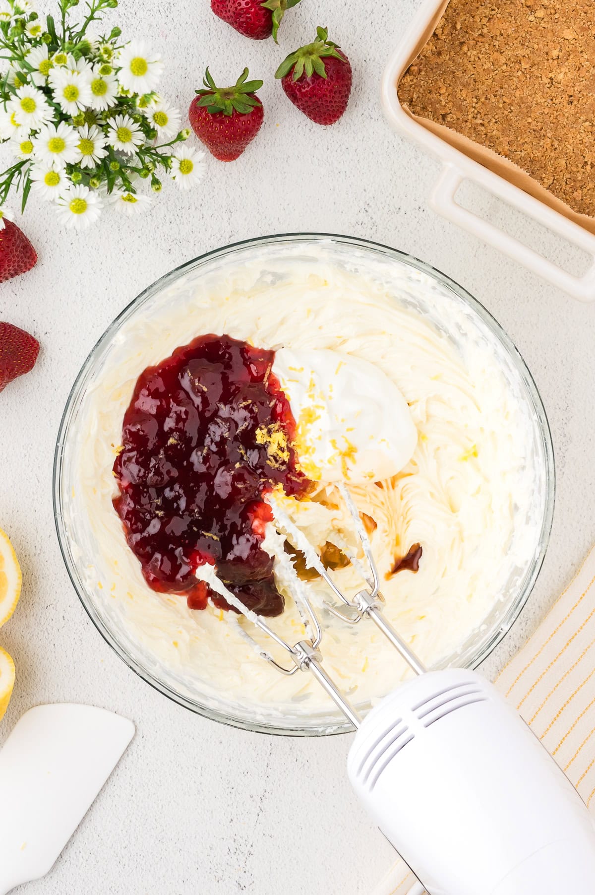 A hand mixer blending cream, jam, and other ingredients in a glass bowl, with strawberries, lemon slices, and a baking dish nearby on a white surface.