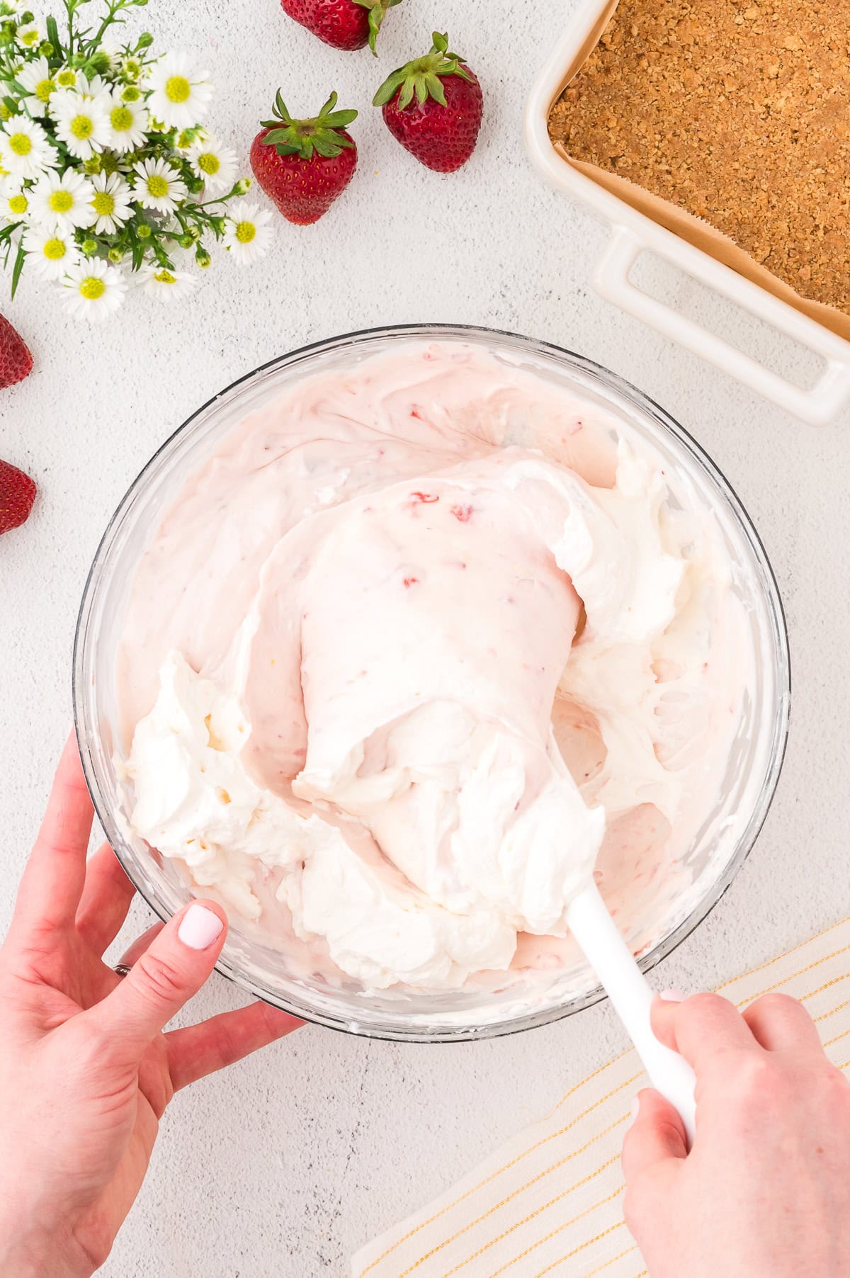 A person mixes a creamy, pink strawberry mixture in a glass bowl with a spatula. Nearby are strawberries, a dish of crumb crust, and a small bouquet of daisies.