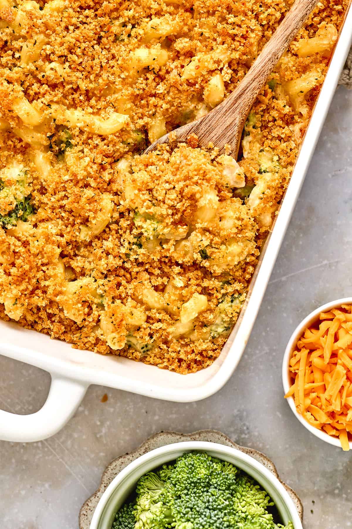 A baking dish of macaroni and cheese with a breadcrumb topping, partially scooped with a wooden spoon, next to bowls of shredded cheese and broccoli.