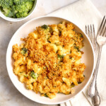 A bowl of broccoli macaroni and cheese topped with breadcrumbs, placed on a napkin with two forks, with a bowl of raw broccoli and a bowl of shredded cheese nearby.